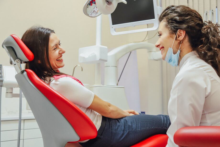 A happy woman with brown hair sitting in a dentists chair smiling & laughing with a female dental hygienist.