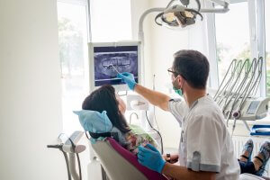 Female patient is being seen by male dentist who is pointing at an x-ray as she looks on.