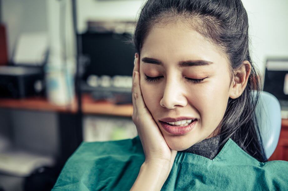 Photograph of a woman holding the right-side of her mouth in pain while in a dentists office.